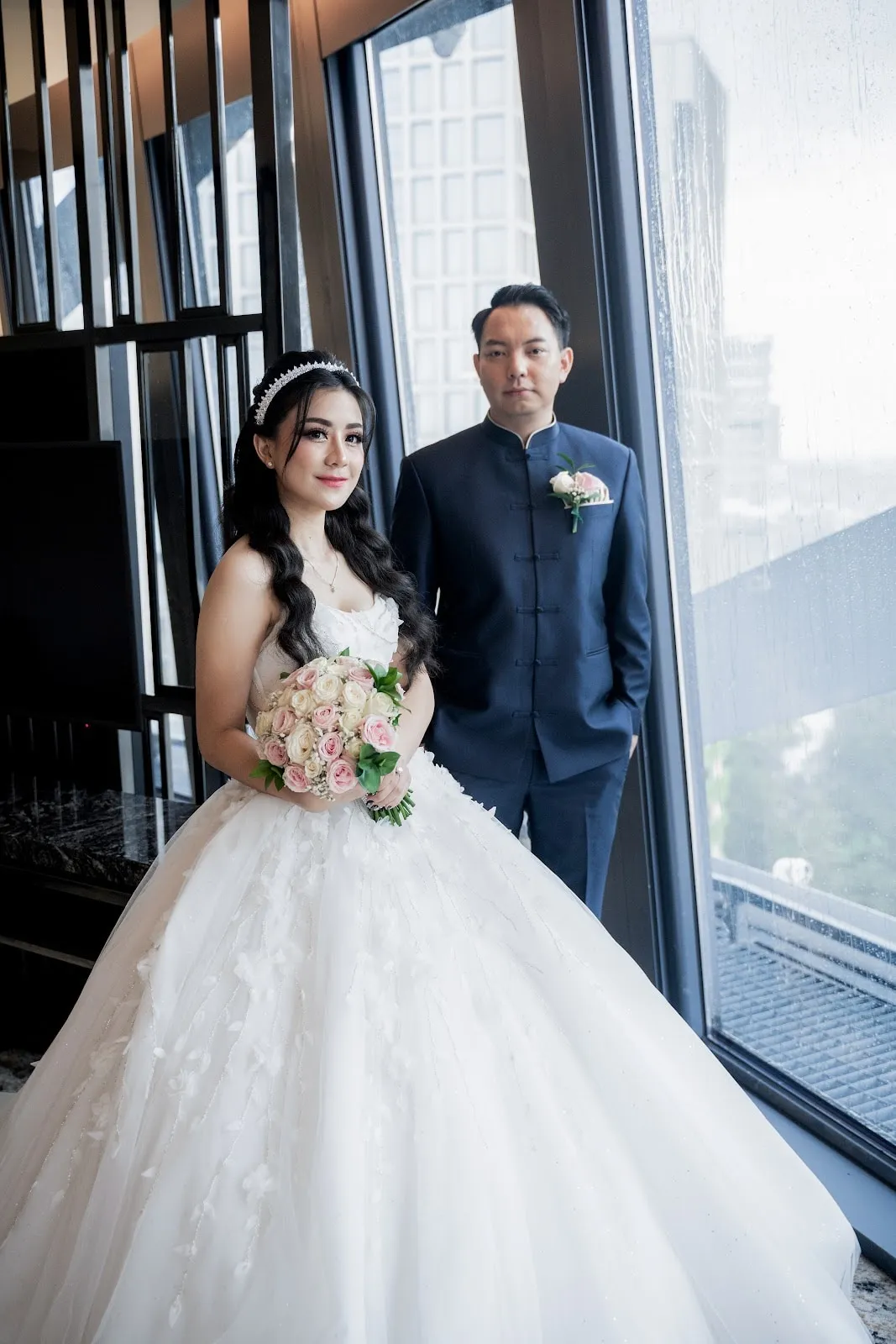 Pasangan pengantin berdiri di tepi jendela dengan buket bunga