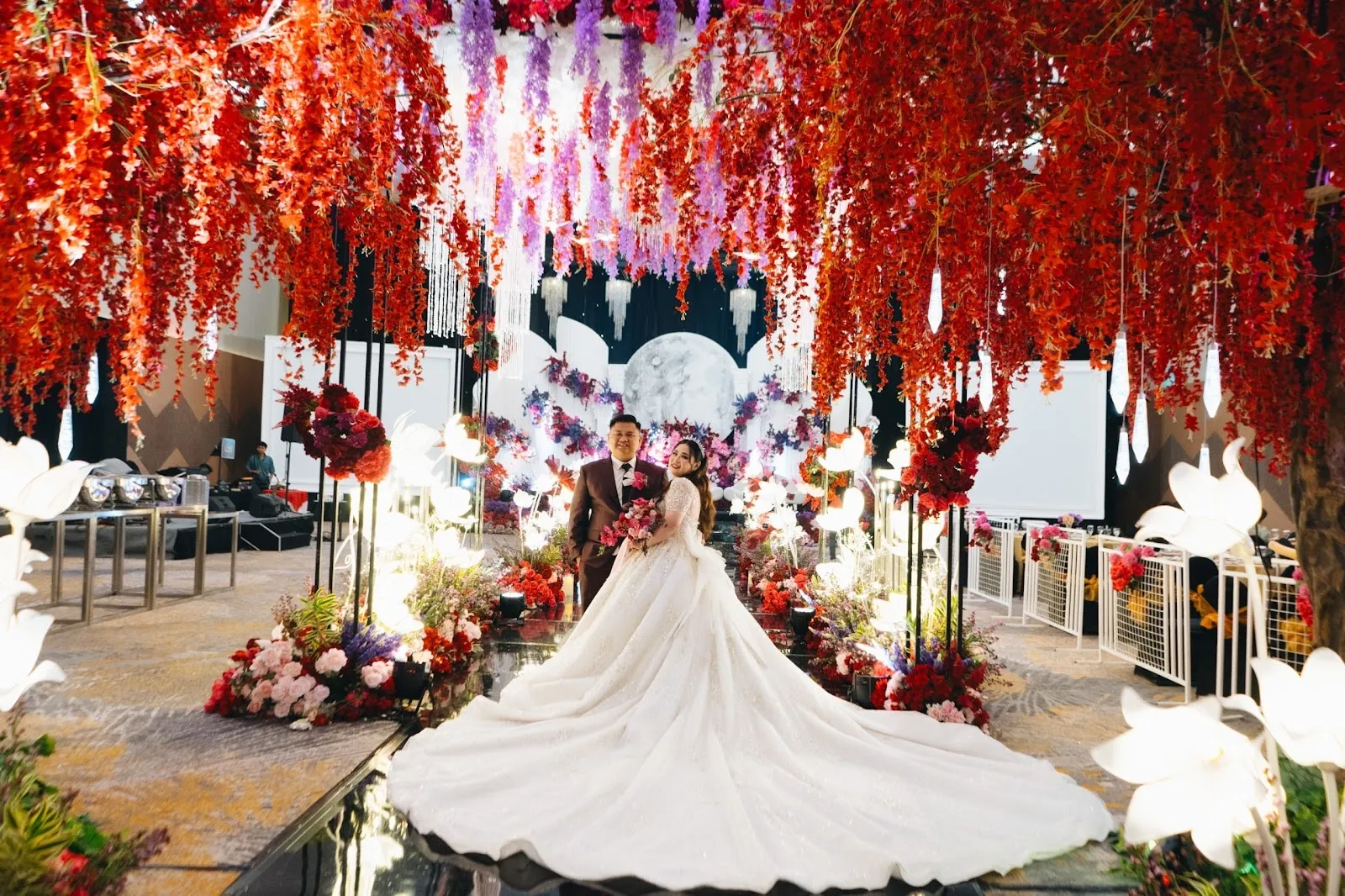Pasangan pengantin di lorong berhias bunga merah dan ungu, gaun berekor sangat panjang